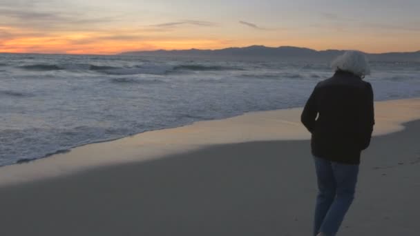 Mujer soltera madura de 60 años con pelo gris caminando por la playa al atardecer — Vídeos de Stock