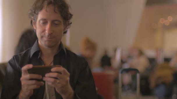 Aantrekkelijke man spelen van een videospel app op horizontale celtelefoon in luchthaven — Stockvideo