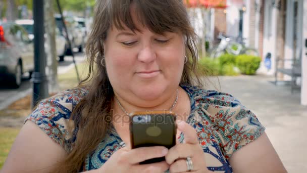 Большая милая кривая женщина в приложении для смс на смартфоне и улыбающаяся на тротуаре — стоковое видео