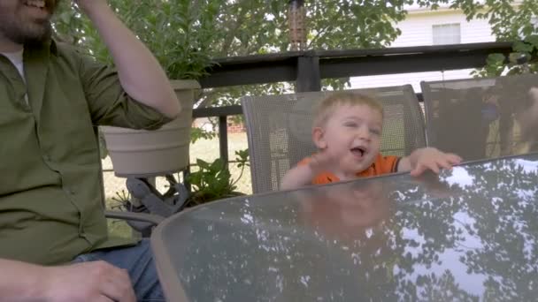 Маленький хлопчик сміється, посміхається і чіпляється з батьком, сидячи за столом — стокове відео
