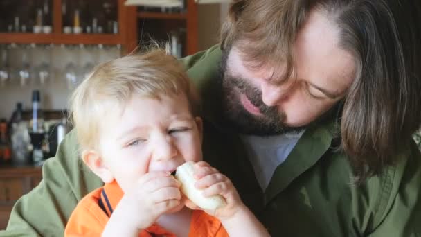 A little boy eats a banana with both hands while his happy father watches — Stock Video