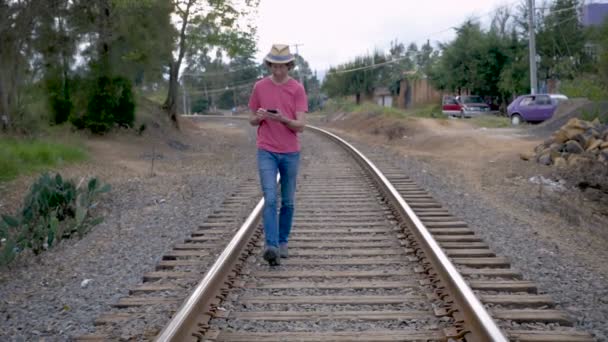 男子戴着帽子走在铁轨上 而低头看着和使用他的智能手机慢动作 — 图库视频影像