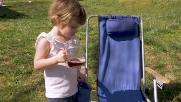 Cute young girl holding a glass mug with juice outside next to a lawn chair — Stock Video