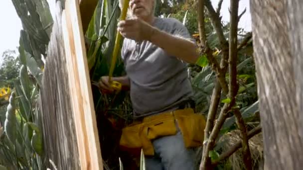 Ángulo bajo de un hombre adulto mayor de 70 años midiendo para una reparación de una cerca casera — Vídeo de stock