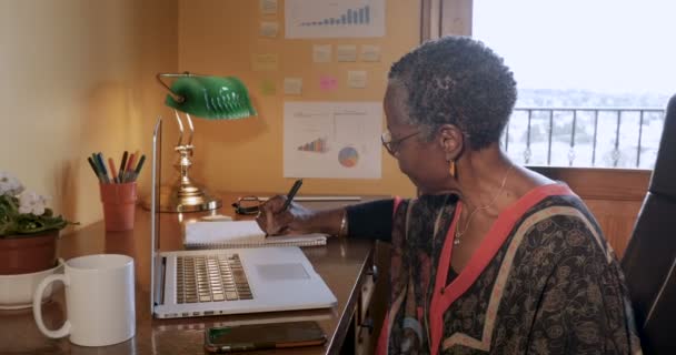 Rijpe zwarte vrouw schrijven in een notitieblok in haar huis of externe kantoor — Stockvideo