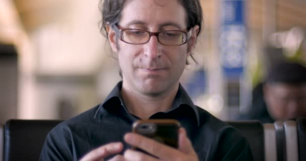 Homem sentado em uma cadeira em um espaço público interior sorrindo em seu telefone — Vídeo de Stock