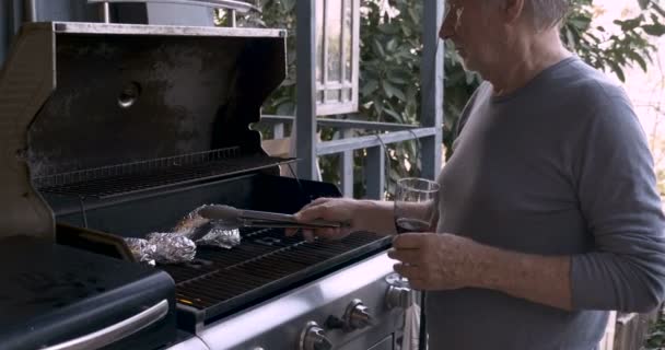 Elderly senior man drinking red wine turning potatoes on a bbq — Stock Video