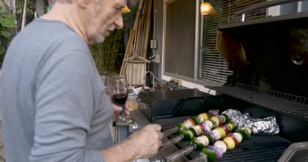 Attractive elderly senior man drinking red wine and grilling vegetable kebabs — Stock Video