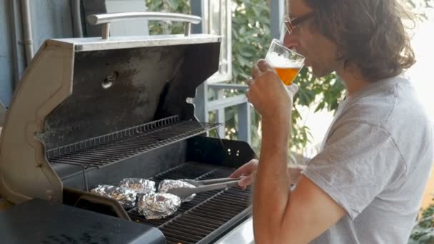 Beau jeune homme buvant une bière transformant des pommes de terre cuites au four sur un barbecue — Video