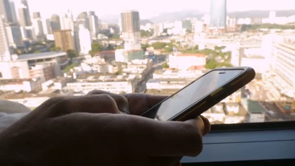 Hombre escribiendo con sus pulgares en un teléfono inteligente con vistas a una ciudad — Vídeos de Stock