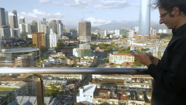Tryck in av en ung man att skriva på sin smarta telefon på en takterrass med utsikt över en stad — Stockvideo