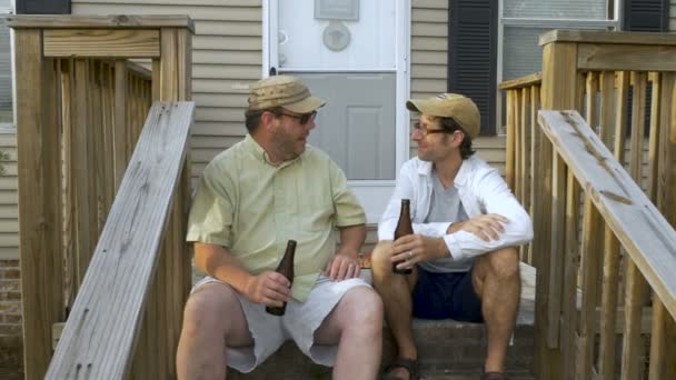 Двое мужчин сидят на крыльце, разговаривают, пьют и расслабляются. — стоковое видео