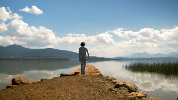 Egy ember séta a mólón, kilátással a hegyi tó szélére — Stock videók