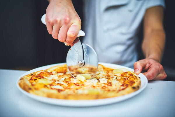 Closeup hand of chef baker in white uniform cutting pizza at kitchen.