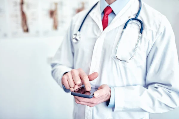 Lääkäri valkoisessa lääketieteellisessä takissa käyttää älypuhelinta ja hymyilee — kuvapankkivalokuva