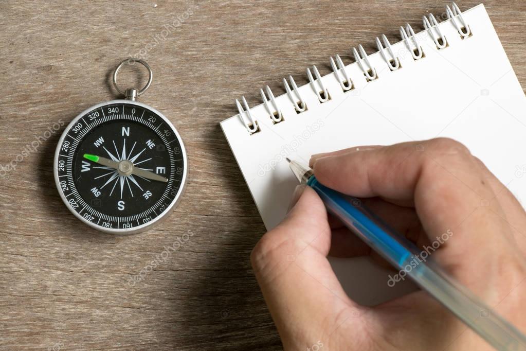 Man hold the pen to write on blank note book with compass on wood desk background