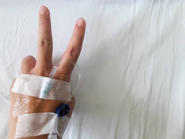 Patient hand with V sign and the tube of normal saline infusion on white cloth background (Victory for the disease) — Stock Photo, Image