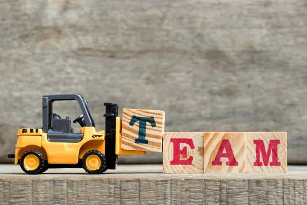 Yellow plastic forklift hold letter T to complete word TEAM on wood background — Stock Photo, Image