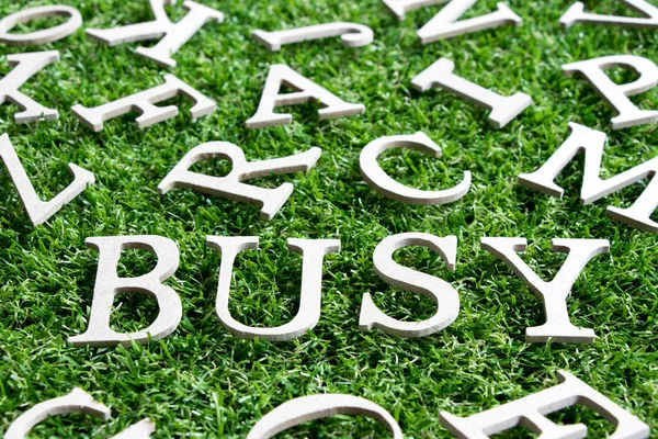 Dřevěná abeceda ve znění zaneprázdněn na umělé trávě zelené pozadí — Stock fotografie