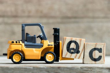Oyuncak sarı forklift ahşap arka plan üzerinde kelime Qc (kalite kontrol kısaltması) tamamlamak için mektup blok Q tutun
