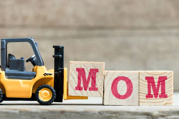 Περονοφόρα toy κίτρινο κρατήστε πατημένο το γράμμα M μπλοκ να ολοκληρώσει τη λέξη μαμά σε ξύλο φόντο (έννοια για την ημέρα της μητέρας) — Φωτογραφία Αρχείου