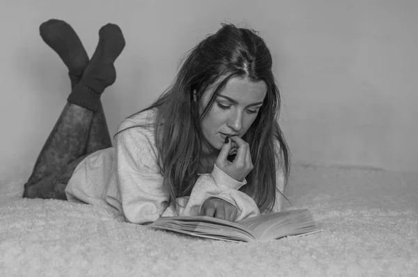 Jovem estudante encantadora com cabelos longos deitado na cama lendo um livro e se preparando para as aulas — Fotografia de Stock