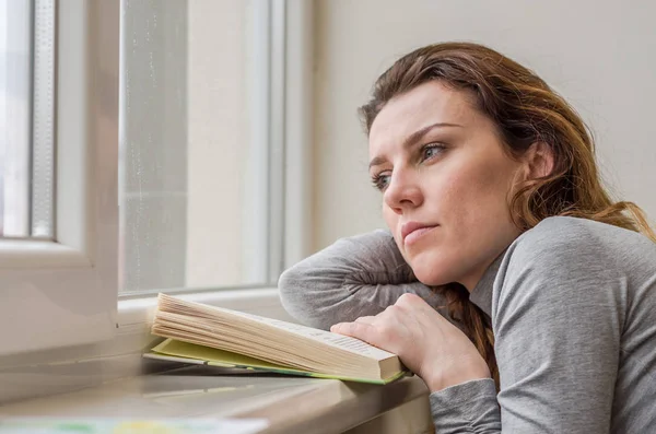 Ung charmig flicka student, med långt hår, ledsen på fönstret läsa bok med lektioner — Stockfoto