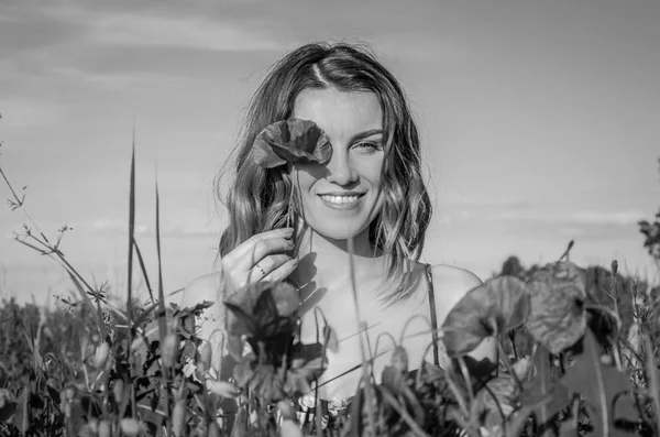 Una joven encantadora en un campo de amapola cierra un ojo con una flor de amapola en un día soleado de verano —  Fotos de Stock