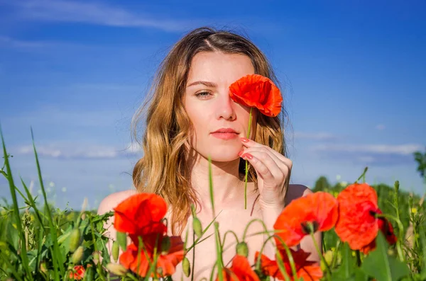 A young charming girl in a poppy field closes one eye with a poppy flower on a bright sunny summer day — Stock Photo, Image