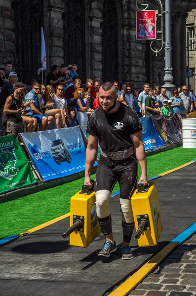 LVIV, UCRANIA - AGOSTO 2017: Un atleta fuerte un culturista lleva maletas pesadas de hierro, en el ejercicio de un paseo del agricultor, en el juego de los hombres fuertes en frente de espectadores entusiastas — Foto de Stock