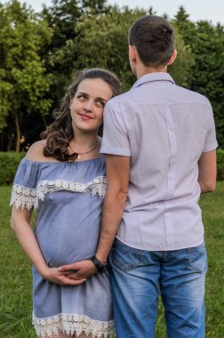 Genç bir çift parkta yürürken, koca hamile karısına karnından sarılır, mutlu bir aile.