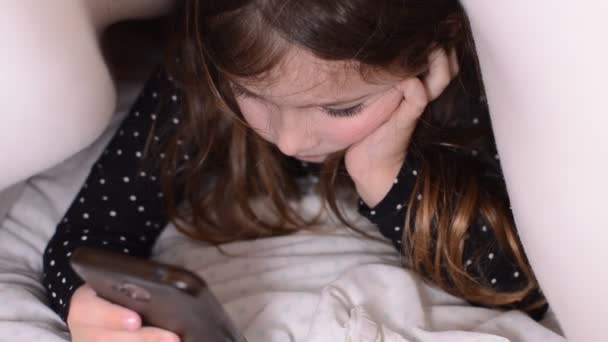 Μικρό Γοητευτικό Κορίτσι Παίζει Ένα Κινητό Τηλέφωνο Κάτω Από Σκεπάσματα — Αρχείο Βίντεο