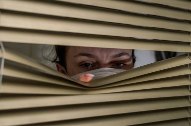 Tıbbi maskeli genç bir kadın pencerenin perdelerinden dışarı bakıyor.