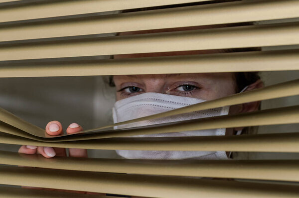 Молодая женщина в медицинской маске смотрит в окно через жалюзи