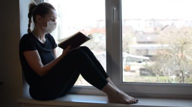 Tıbbi maskeli genç bir kız pencerenin kenarında oturur ve kitap okur.