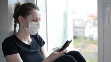 Tıbbi maskeli genç bir kız pencerenin kenarındaki pencere eşiğinde oturuyor ve akıllı telefona bakıyor.