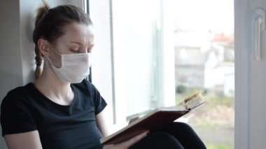 Tıbbi maskeli genç bir kız pencerenin kenarında oturur ve kitap okur.