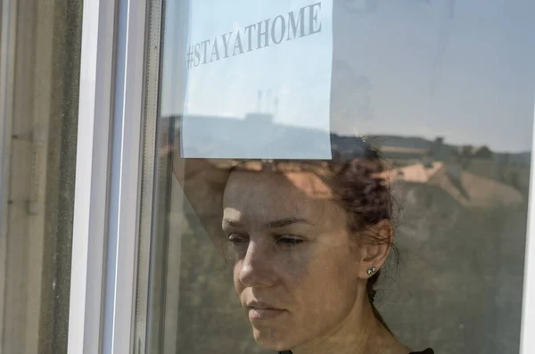 A young girl stands by the window with a sign saying stayathome