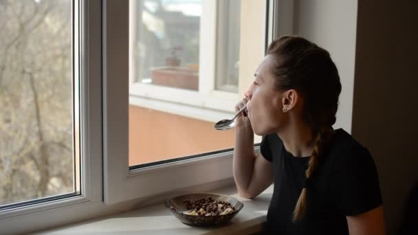 창가에 여자가 요구르트를 곁들인 초콜릿 곡류를 먹는다 — 비디오