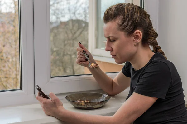 窗户旁边的年轻女子一边吃着含酸奶的巧克力早餐谷类食品 一边坐在智能手机前 — 图库照片
