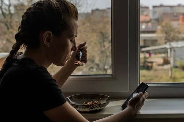 窗户旁边的年轻女子一边吃着含酸奶的巧克力早餐谷类食品 一边坐在智能手机前 — 图库照片