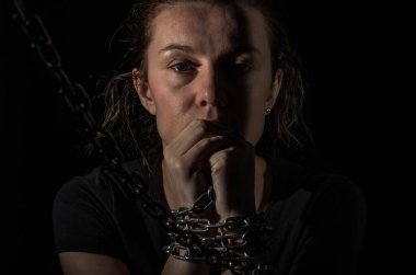 Zincirlerle bağlanmış genç bir kadın siyah bir arka planda ortaya çıkıyor.