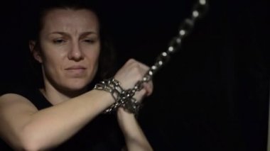 Zincirlerle bağlanmış genç bir kadın siyah bir arka planda ortaya çıkıyor.