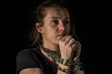 Zincirlerle bağlanmış genç bir kadın siyah bir arka planda ortaya çıkıyor.