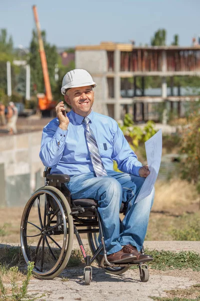 Disabled person in the construction helmet with documents in hand talking on the phone on the background of building. Successful wheelchair user directs the work.