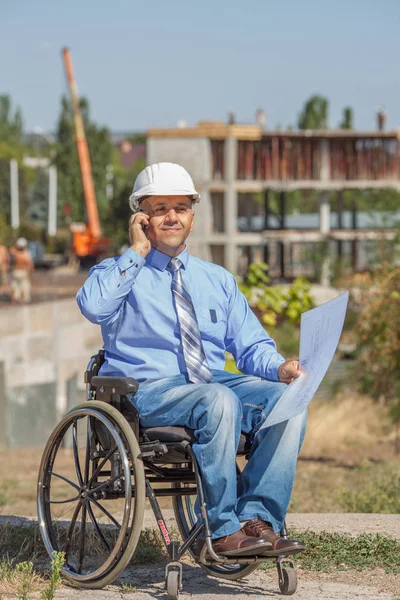 Disabled person in the construction helmet with documents in hand talking on the phone on the background of building. Successful wheelchair user directs the work.