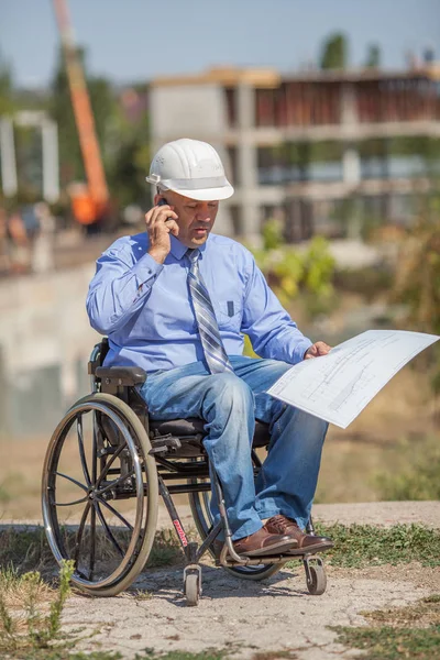 Disabled person in the construction helmet with documents in hand talking on the phone on the background of building. Successful wheelchair user directs the work.