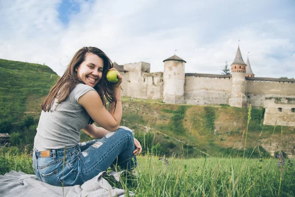 Pretty tourist sits on rock in front of old castle and eat aplle — Stock Photo, Image