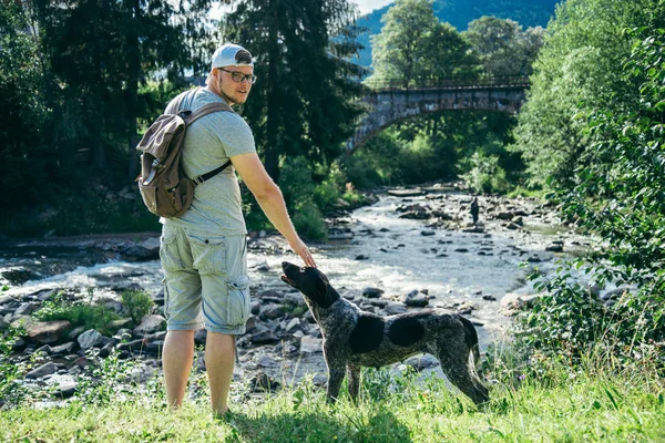 Jovem homem forte fica perto de montanhas rio com cão e à procura da vista — Fotografia de Stock
