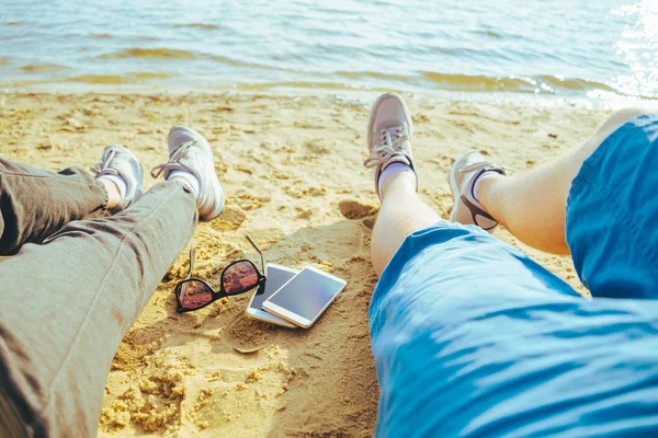 Mans and womans legs on the beach, mobile phones with sunglasses between them — Stock Photo, Image
